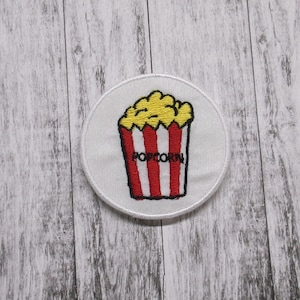 May include: A white round embroidered patch with a red and white striped popcorn bucket filled with yellow popcorn. The bucket has the word "POPCORN" written on it.