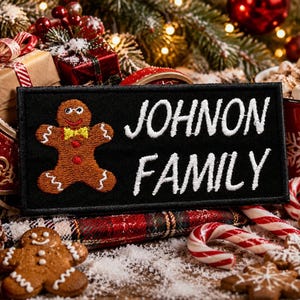 May include: A black rectangular patch embroidered with a gingerbread man and the words "JOHNSON FAMILY" in white. The patch is surrounded by Christmas decorations, including gifts, candy canes, and gingerbread cookies.