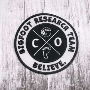 Puede incluir: Un parche bordado blanco y negro con el texto "Bigfoot Research Team Believe" en un círculo alrededor de un gráfico en blanco y negro de una huella y las letras "CO" con una cruz.