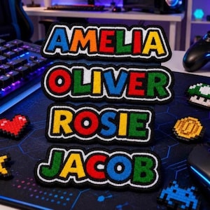 Könnte beinhalten: Vier rechteckige bestickte Aufnäher mit den Namen AMELIA, OLIVER, ROSIE und JACOB in einer kräftigen, mehrfarbigen Pixel-Schriftart. Jeder Buchstabe ist schwarz umrandet, wobei die Namen in leuchtendem Rot, Blau, Gelb und Grün dargestellt sind.