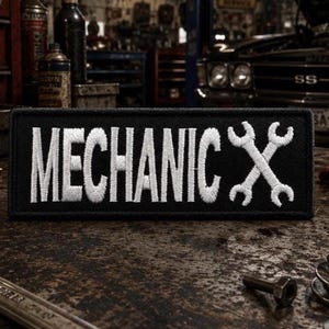 May include: A rectangular black embroidered patch featuring the white text MECHANIC in a bold font followed by a graphic of two crossed white wrenches. The patch has a clean stitched border and is designed for attachment to clothing or gear.
