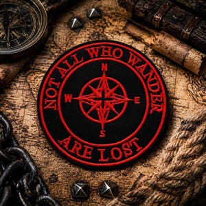 May include: A black circular patch with red embroidered text and a compass rose design. The text reads "NOT ALL WHO WANDER ARE LOST". The patch is set against a backdrop of a vintage map, a compass, and other nautical-themed items.