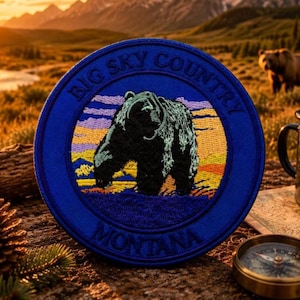 Może przedstawiać: Okrągła, niebieska naszywka z napisem "BIG SKY COUNTRY MONTANA" otaczającym szczegółową ilustrację czarnego niedźwiedzia. Niedźwiedź jest czarny, na tle zachodzącego słońca. Widoczny jest również czarny kubek emaliowany i kompas.