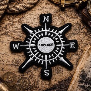 Könnte beinhalten: Ein schwarz-weiß bestickter Aufnäher in Form einer Kompassrose. Das Design zeigt einen zentralen Kreis mit dem in Schwarz gestickten Wort EXPLORE, umgeben von den Himmelsrichtungsbuchstaben N, S, E und W an den Spitzen der Kompassnadeln.