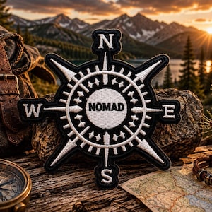 May include: A black and white embroidered patch shaped like a compass rose with the word NOMAD printed in the center. The patch features cardinal directions N, S, E, and W at the points of the compass, with detailed stitching throughout the design.