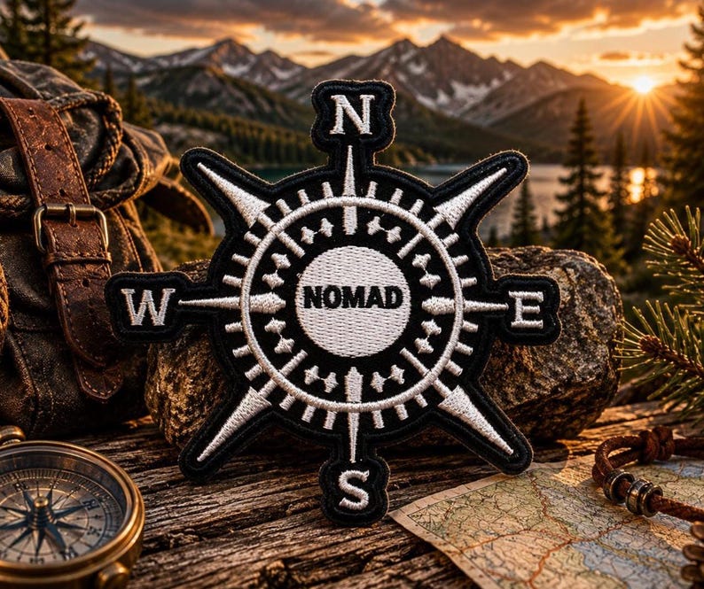 May include: A black and white embroidered patch shaped like a compass rose with the word NOMAD printed in the center. The patch features cardinal directions N, S, E, and W at the points of the compass, with detailed stitching throughout the design.