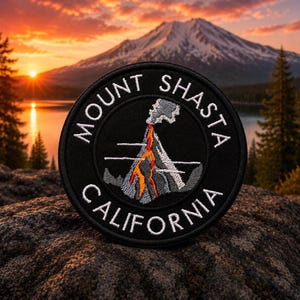 Może przedstawiać: Czarna okrągła naszywka z białym haftowanym napisem "MOUNT SHASTA CALIFORNIA". Naszywka przedstawia haftowany obraz wybuchającego wulkanu ze szczegółami w kolorze pomarańczowym i szarym. Tło przedstawia górę, jezioro i zachód słońca.
