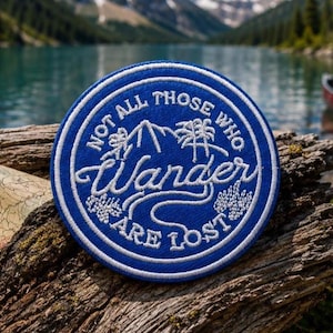 May include: A circular embroidered patch featuring a deep blue background with white stitching. The design includes a mountain peak, pine trees, and the text "NOT ALL THOSE WHO WANDER ARE LOST" in a stylised font, all enclosed within a white border.