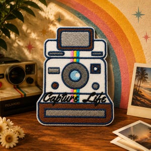 May include: Embroidered patch in the shape of a vintage camera with the words "Capture Life." The patch is white with blue, brown, pink, and yellow accents. A retro rainbow and a Polaroid camera are in the background.