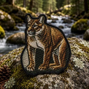 May include: An embroidered patch featuring a mountain lion in shades of brown, tan, and black. The feline is depicted in a standing profile position with detailed fur texture and a dark stitched border, showcasing a realistic animal design.