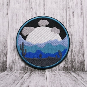 May include: Embroidered patch with a desert landscape scene. The patch features a large white moon, blue mountains, and gray clouds against a black background. There are small green cacti in the foreground.