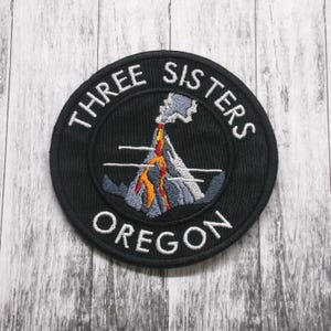 May include: A black embroidered patch with the text "Three Sisters Oregon" and a stylized image of a volcano with smoke and lava.