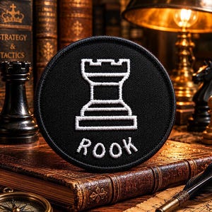 May include: A black circular patch featuring a white embroidered rook chess piece and the word "ROOK". The patch is set against a backdrop of antique books and chess pieces, suggesting a theme of strategy and tactics.