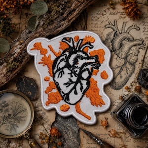 May include: An embroidered patch featuring a black anatomical heart outline centered on a white background with irregular orange splatters. The patch has a white border and an organic, non-uniform shape, showcasing detailed black thread work.