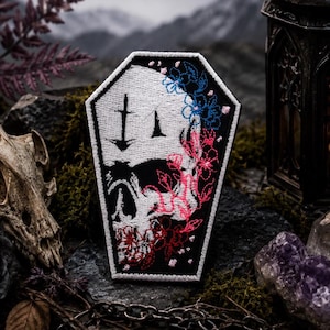 Könnte beinhalten: Ein sargförmiger Stickaufnäher mit einem weißen Totenkopfmotiv auf schwarzem Hintergrund. Der Totenkopf ist mit blauen, rosafarbenen und roten Blumenstickereien an der rechten Seite sowie einem kleinen schwarzen Schwertsymbol auf der Stirn verziert.