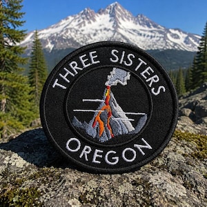 Może przedstawiać: Czarna okrągła naszywka z białym haftowanym napisem „THREE SISTERS OREGON”. Naszywka przedstawia pasmo górskie z wulkanem wyrzucającym pomarańczowy i szary dym. Tło przedstawia pasmo górskie i drzewa.