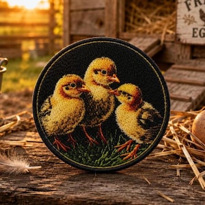 May include: A circular embroidered patch featuring three yellow chicks standing on green grass against a black background. The embroidery depicts the chicks with detailed golden-yellow feathers, orange beaks, and small feet, all enclosed within a stitched black border.