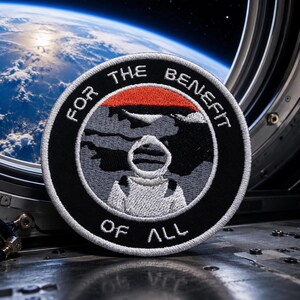 May include: Circular embroidered patch with the text "FOR THE BENEFIT OF ALL" around the edge. The patch features an astronaut in a spacesuit, a red stripe, and a gray background, set against a view of Earth from space.