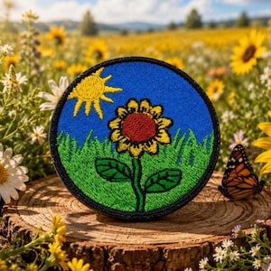 Puede incluir: Parche bordado redondo con un girasol con centro rojo, pétalos amarillos y tallo verde, sobre un cielo azul con un sol amarillo. El parche tiene un borde negro y está sobre una superficie de madera.