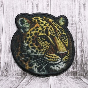 Pode incluir: Close-up de um remendo bordado circular que apresenta uma imagem detalhada do rosto de um leopardo. O leopardo tem o nariz preto, os olhos amarelos e uma pelagem manchada de preto e marrom.