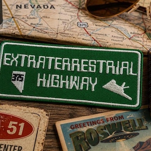Może przedstawiać: Prostokątna, zielona haftowana naszywka z białym napisem EXTRATERRESTRIAL HIGHWAY. Naszywka zawiera biały zarys stanu Nevada z liczbą 375 w środku oraz białą grafikę latającego spodka po prawej stronie.