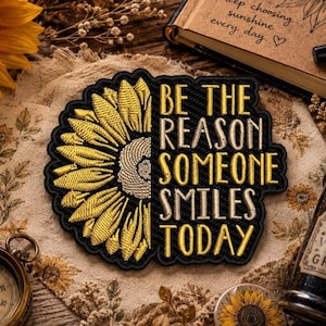 May include: Embroidered patch featuring a sunflower design with the text "BE THE REASON SOMEONE SMILES TODAY." The patch has a black border and yellow and gold details. The patch is on a beige floral background.