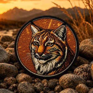 May include: A circular embroidered patch featuring a detailed portrait of a bobcat with orange, brown, and white fur. The cat has pointed ears, amber eyes, and long whiskers. The patch is framed by a thick black border and has a textured finish.