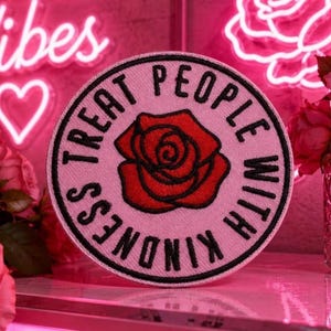 May include: A round pink patch with the words "TREAT PEOPLE WITH KINDNESS" encircling a red rose. The patch is on a pink surface, with a neon sign that says "vibes" in the background.