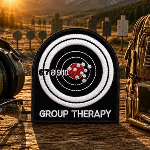 May include: A black embroidered patch featuring a white circular target design with the numbers 6, 7, 8, 9, and 10 printed in white. The center of the target is red with white bullet hole graphics. The text GROUP THERAPY is embroidered in white.