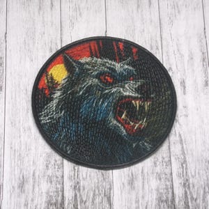 Może przedstawiać: Okrągła naszywka przedstawiająca warczącego wilkołaka z czerwonymi oczami i ostrymi zębami. Projekt zawiera czerwony i pomarańczowy zachód słońca z żółtym księżycem i sylwetkami drzew. Naszywka ma czarną obwódkę.