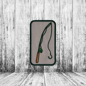 May include: A rectangular patch featuring a fishing rod design. The patch has a light gray background with a dark green border. The fishing rod is depicted in green with an orange handle and a green bobber.