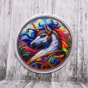 Puede incluir: Un parche bordado colorido con un unicornio blanco con una crin y un cuerno arcoíris. El unicornio está rodeado de diseños coloridos y en espiral.