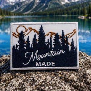 May include: A rectangular embroidered patch featuring a white background with a dark blue silhouette of a forest and a gold-coloured mountain outline. The text Mountain MADE is embroidered in white script and block letters across the lower section.