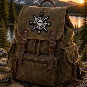 May include: An olive green canvas backpack featuring brown leather straps with metal buckles, a drawstring closure, and side pockets. A black and white circular patch on the top flap displays a compass design with the text NOMAD in the center.