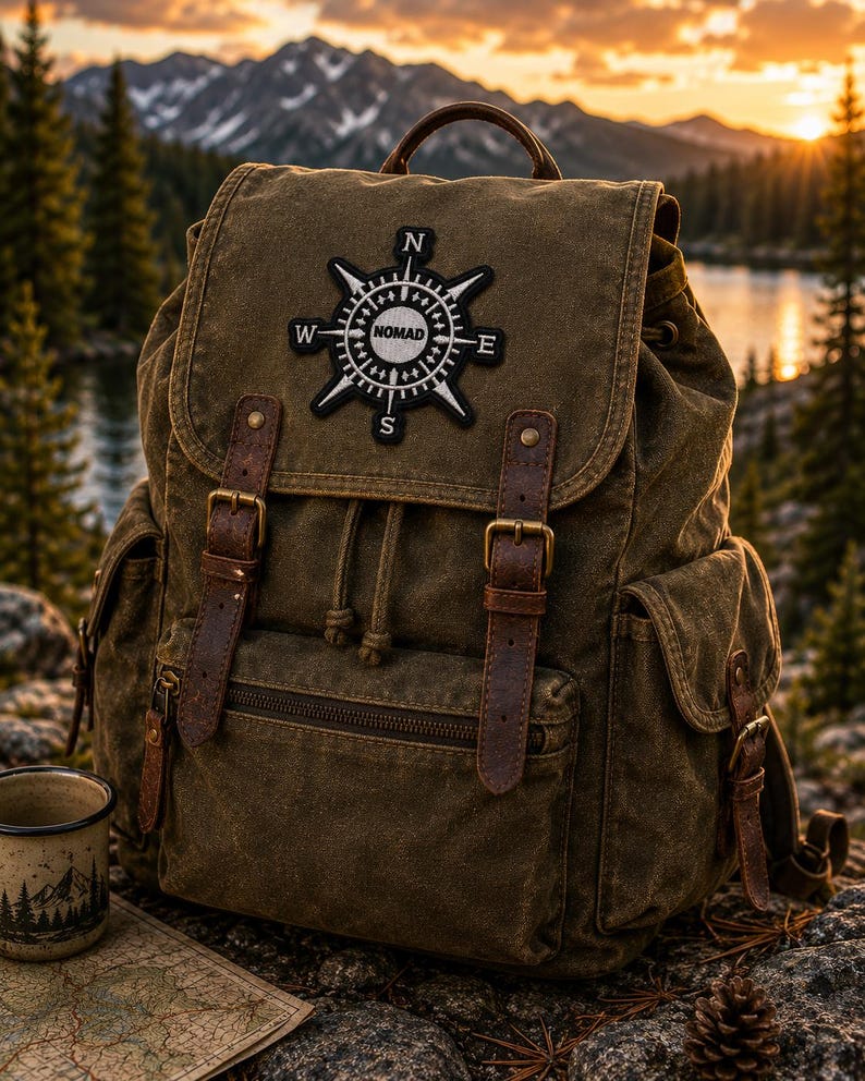 May include: An olive green canvas backpack featuring brown leather straps with metal buckles, a drawstring closure, and side pockets. A black and white circular patch on the top flap displays a compass design with the text NOMAD in the center.