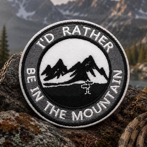 Peut inclure: Un écusson brodé circulaire présentant un paysage de montagne en noir et blanc. La bordure contient le texte blanc I'D RATHER BE IN THE MOUNTAINS. Une petite icône de panneau de signalisation blanc est située à la base des montagnes.