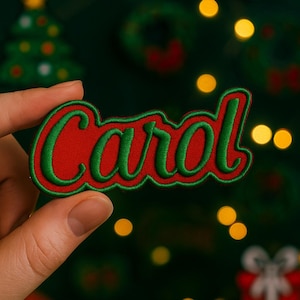 Puede incluir: Un parche bordado rojo con el nombre "Carol" en escritura verde. El parche tiene un borde verde y se sostiene contra un fondo borroso de decoraciones y luces navideñas. El parche es un artículo festivo.