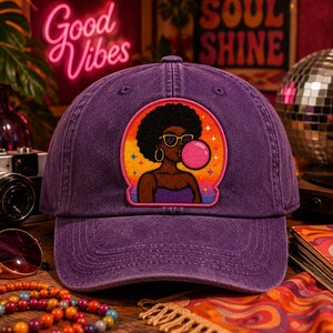 Pode incluir: Um bon&eacute; de beisebol roxo com um emblema bordado central. O emblema apresenta uma ilustra&ccedil;&atilde;o gr&aacute;fica de uma pessoa com penteado afro, usando &oacute;culos de sol amarelos e brincos de argola dourados, soprando uma grande bolha de chiclete rosa.