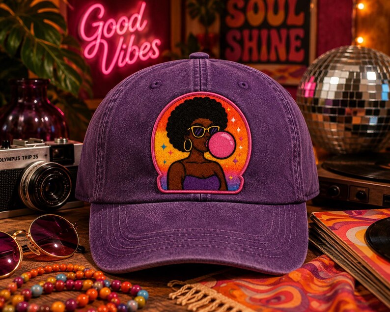 Pode incluir: Um bon&eacute; de beisebol roxo com um emblema bordado central. O emblema apresenta uma ilustra&ccedil;&atilde;o gr&aacute;fica de uma pessoa com penteado afro, usando &oacute;culos de sol amarelos e brincos de argola dourados, soprando uma grande bolha de chiclete rosa.