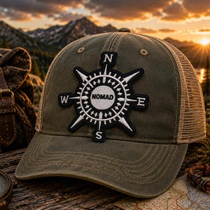 May include: An olive green baseball cap with a mesh back and a black and white embroidered patch on the front. The patch features a compass design with the cardinal directions N, E, S, W and the word NOMAD printed in the center of the circle.