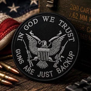May include: A circular black embroidered patch featuring a white eagle emblem at the center with rifles on each side. The text IN GOD WE TRUST is arched across the top, and the text GUNS ARE JUST BACKUP is arched across the bottom.