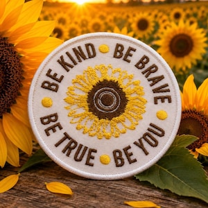 May include: A circular embroidered patch with a white background featuring a central yellow sunflower with a brown center. The text BE KIND, BE BRAVE, BE TRUE, and BE YOU is stitched in brown around the sunflower, separated by small yellow dots.