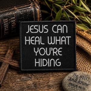 May include: A black embroidered patch with the text "JESUS CAN HEAL WHAT YOU'RE HIDING" in white. A small, gray stone with the inscription "Be still and know PSALM 46:10" is also visible.