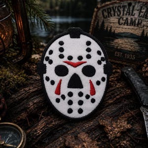 May include: A white hockey mask patch with a black border, featuring black circular ventilation holes, two black eye openings, a black triangular nose opening, and red chevron accents above the eyes and below the nose on a textured fabric surface.