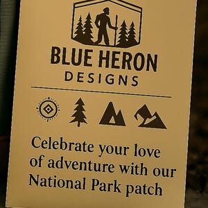 May include: A tan card with the text "BLUE HERON DESIGNS" and a logo of a person hiking in front of trees. The card also says "Celebrate your love of adventure with our National Park patch."