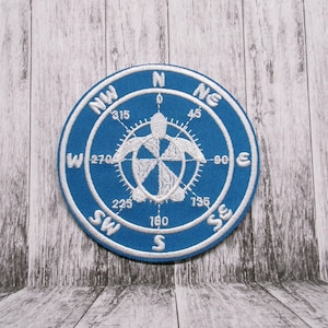 May include: A blue and white embroidered patch with a compass design featuring a sea turtle in the center. The compass points are labeled N, NE, E, SE, S, SW, W, and NW. The numbers 0, 45, 90, 135, 180, 225, 270, and 315 are also included.