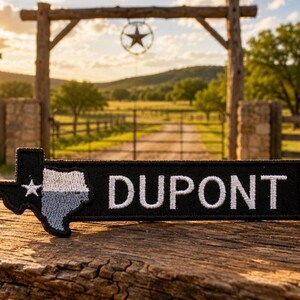 May include: A rectangular black embroidered patch featuring the word DUPONT in white capital letters next to a Texas-shaped emblem. The Texas shape is divided into a white star on a black background and a grey textured section with a black border.