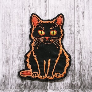 May include: Black embroidered cat patch with orange outline and yellow eyes. The cat is sitting with its paws tucked under its body.