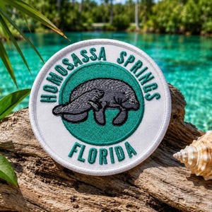 May include: A circular patch featuring the text "HOMOSASSA SPRINGS FLORIDA" around a grey manatee illustration on a teal green background. The patch is white with a stitched border, placed on driftwood.