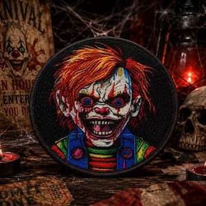 May include: A circular embroidered patch featuring a detailed depiction of a menacing clown with bright orange hair, piercing blue eyes, and a wide, toothy grin. The clown wears a blue vest over a multi-colored striped shirt with red accents.